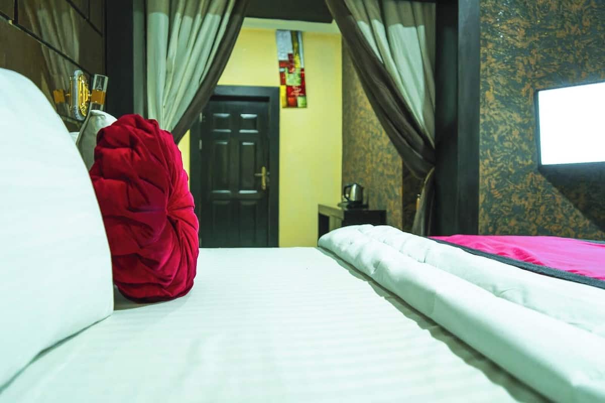 Una habitación de hotel con una cama, una colcha roja, cortinas, una mesita de noche con una lámpara y una ventana con vistas al exterior.