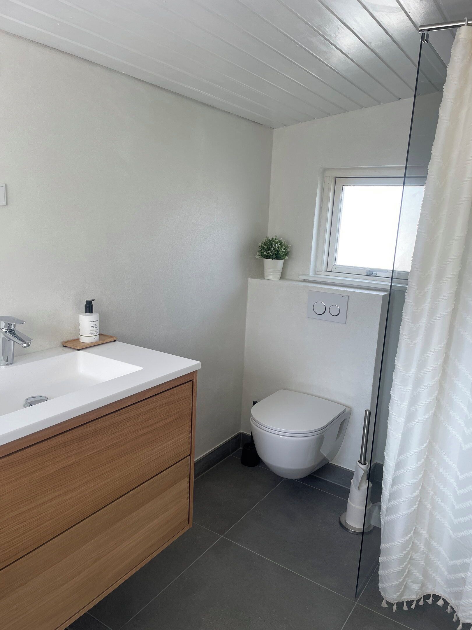 Une salle de bain avec un lavabo blanc, un meuble-lavabo en bois, des toilettes, une petite fenêtre et un rideau de douche.