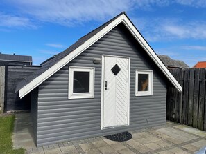 A small, single-story building with a white door and two windows, a sloped roof, and a wooden fence in the background. - Lovely holiday home 80 m from the water (Otterup)