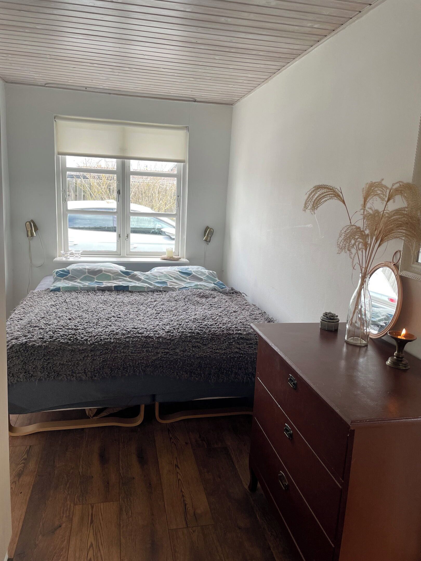A bedroom with a bed, wooden dresser, window, and a vase with dried plants.