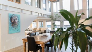A spacious living area with a wooden dining table, comfortable seating, and large windows offering views of the outdoors. - Stylish Art Loft with Spa & Soaring Ceilings (Christchurch)