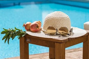 Una mesa de madera junto a una piscina, con un sombrero tejido, gafas de sol y granadas.
