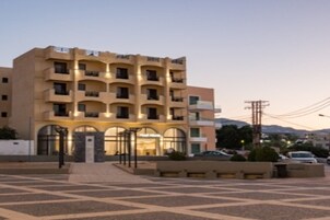 Un hotel de varios pisos con entrada central, balcones en cada planta y un área de estacionamiento al frente.
