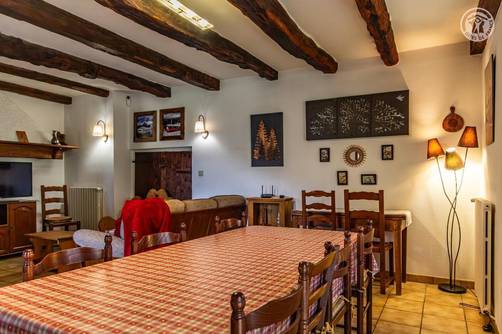 A dining area with a large table, wooden chairs, and a red tablecloth. There are wall-mounted lamps and framed pictures on the walls.