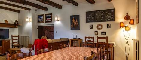 A dining area with a large table, wooden chairs, and a red tablecloth. There are wall-mounted lamps and framed pictures on the walls.