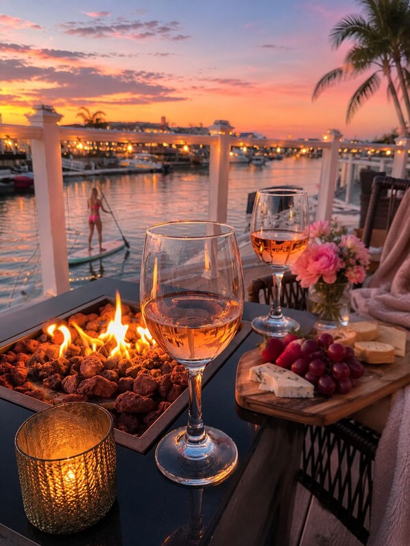 A table set with wine glasses, a tray of snacks, and a candle, overlooking a marina at sunset.