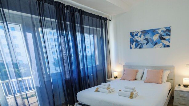 Un dormitorio con cama, mesita de noche, lámpara, un cuadro y vistas al exterior.