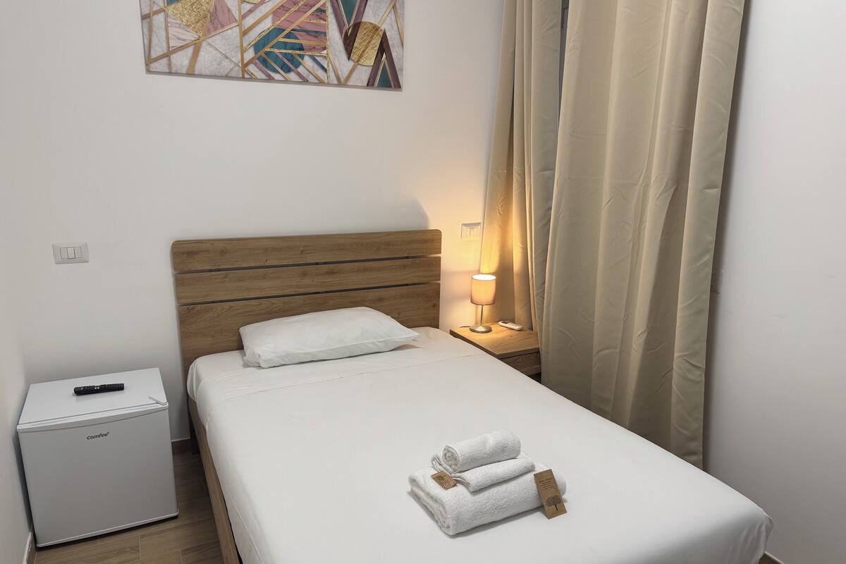 A neatly made bed with white linens and two rolled towels, a bedside table with a lamp and remote control, a small white cabinet, and a wall art piece above the bed.
