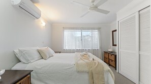 Ein Schlafzimmer mit Bett, Deckenventilator, Fenster mit leichtem Vorhang, Nachttisch und Kleiderschrank.