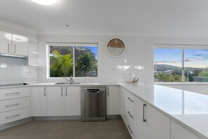 A modern kitchen with white cabinetry, a stainless steel dishwasher, and a large window offering a view of the outdoors. - Sea Sage Coastal Retreat in Fingal Bay (Fingal Bay)