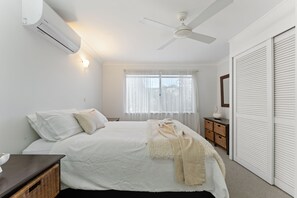 A bedroom with a bed, a ceiling fan, a window with curtains, a bedside table, and a wardrobe. - Sea Sage Coastal Retreat in Fingal Bay (Fingal Bay)