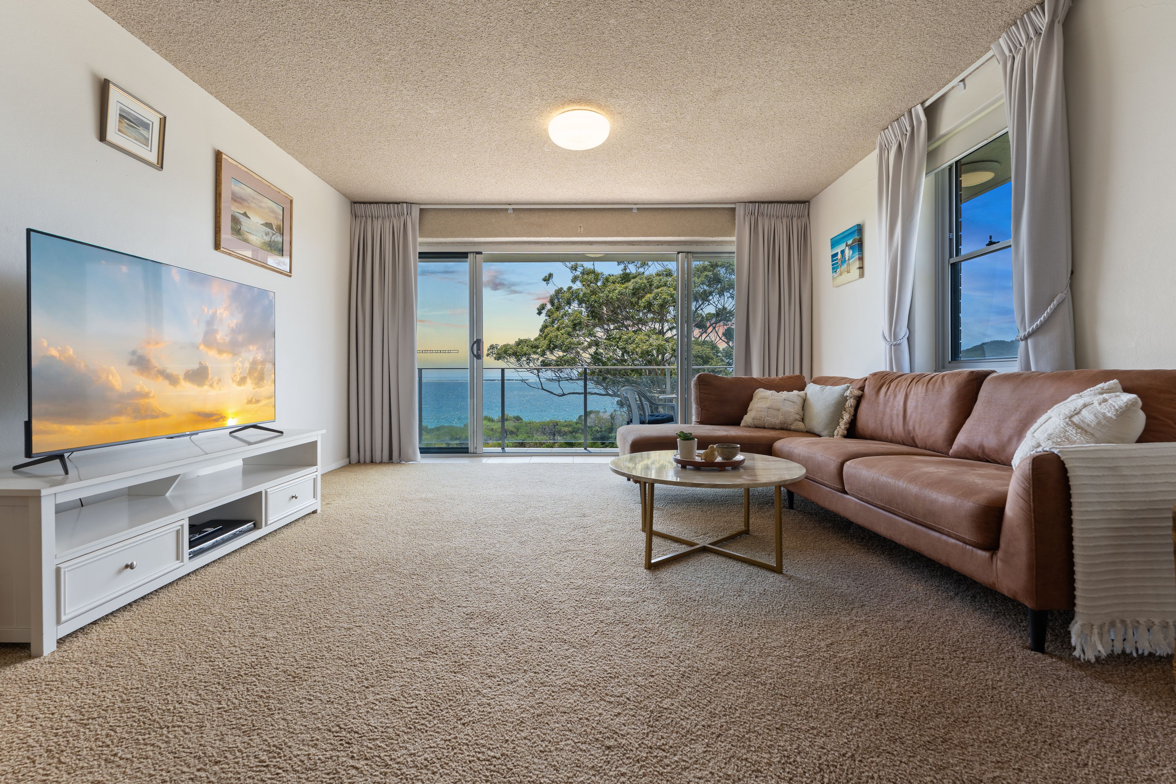 A living room with a flat-screen TV, a brown leather sofa, a coffee table, and a large window with a view of the sea.