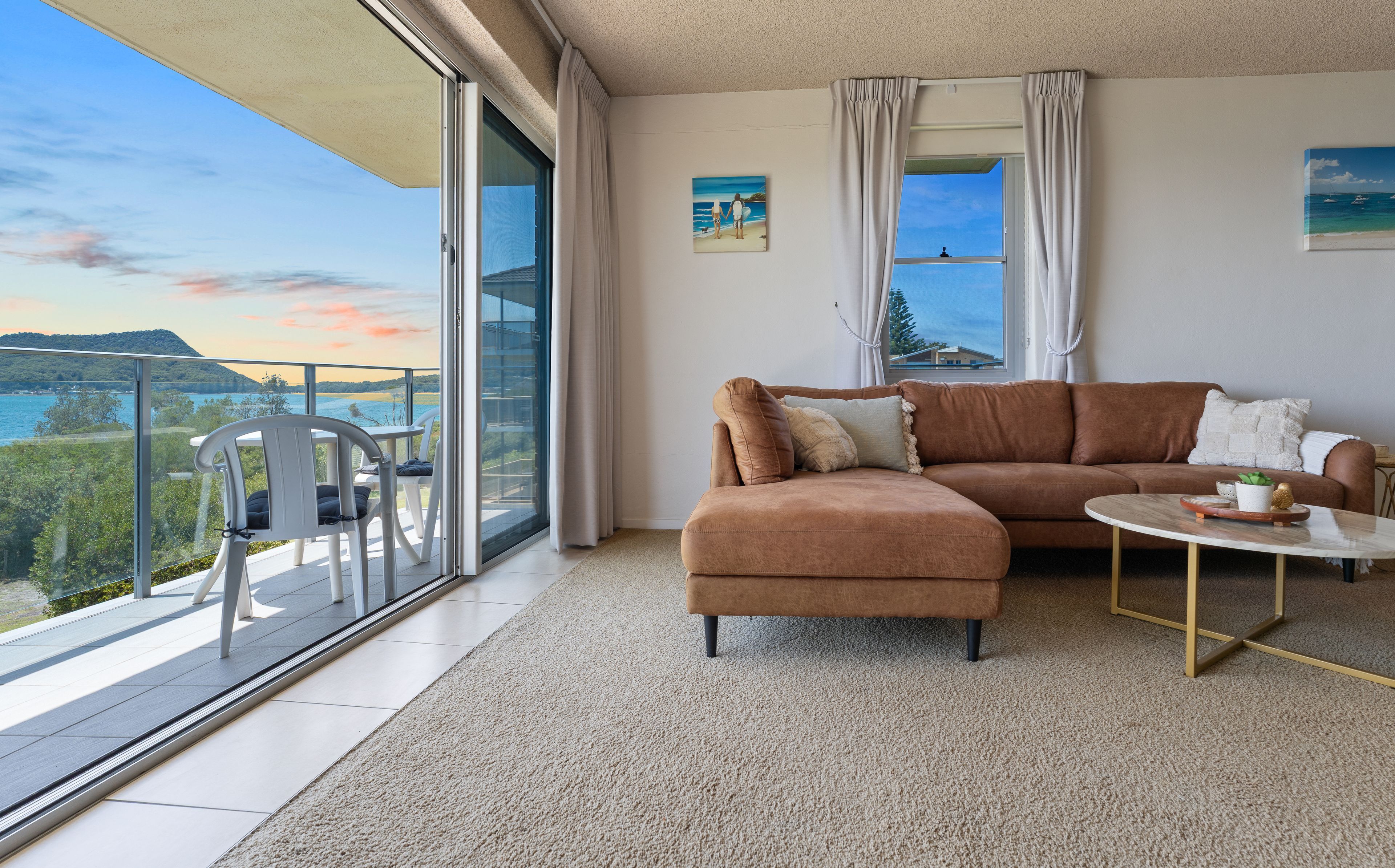 A modern living room with a brown sofa, a small round table, and a balcony with a view of the sea and hills.