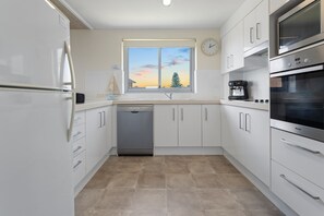 A modern kitchen with white cabinetry, a dishwasher, a coffee machine, and a clock on the wall. - Shoal Bay Sunrise Waterfront Apartment Nelson Bay (Nelson Bay)