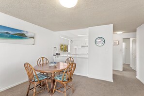 A dining area with a round table and four chairs, a kitchen in the background, and a clock on the wall. - Shoal Bay Sunrise Waterfront Apartment Nelson Bay (Nelson Bay)