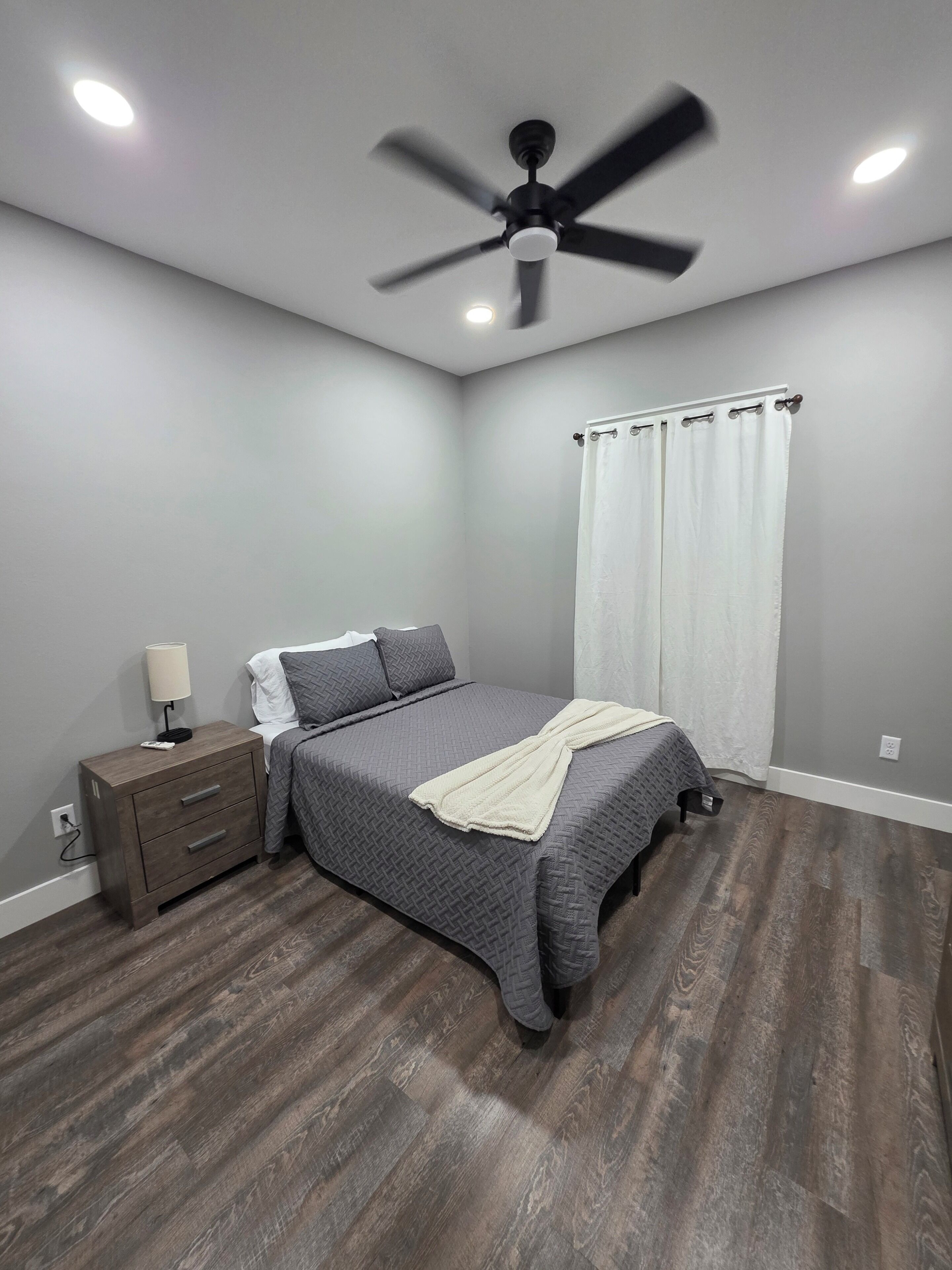 Un dormitorio con cama, mesita de noche, lámpara, ventilador de techo y una ventana con cortinas.