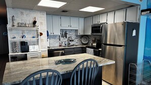 A modern kitchen with white cabinets, a stainless steel refrigerator, and a marble island with two chairs. - Beachfront condo w/ balcony in wonderful Garden City Beach (Murrells Inlet)