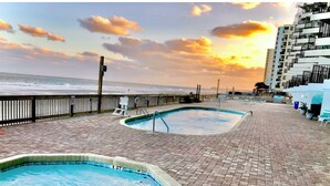A pool area with two pools, a wooden deck, and a view of the ocean at sunset. - Beachfront condo w/ balcony in wonderful Garden City Beach (Murrells Inlet)
