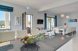 A modern dining area with a central island featuring a fruit bowl, a dining table set for six, and a view of the outdoors through large windows. - Villa Rachele by Istrabook (Rakalj)