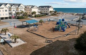 A playground with a blue slide and climbing structures, surrounded by a sandy area with palm trees. - Otter Oasis - Serene 3bd pet-friendly sound views (Surf City)