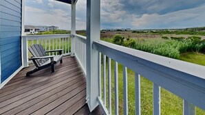 A wooden deck with a white railing, a single Adirondack chair, and a view of a grassy landscape with buildings in the distance. - Otter Oasis - Serene 3bd pet-friendly sound views (Surf City)