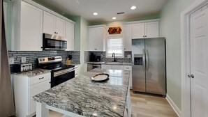 A modern kitchen with white cabinets, a granite countertop, stainless steel appliances, and a window with blinds. - Otter Oasis - Serene 3bd pet-friendly sound views (Surf City)