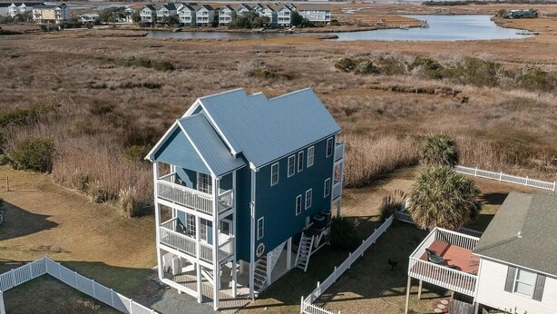 A two-story house with a balcony, surrounded by a white picket fence, set in a grassy area with a body of water and other houses in the background. - Otter Oasis - Serene 3bd pet-friendly sound views (Surf City)
