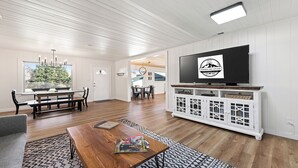 Un vaste salon doté d’un plancher en bois, un coin repas avec une table et des chaises, un téléviseur fixé sur un meuble blanc et un lustre.