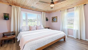 Une chambre avec un grand lit, un plancher en bois, un ventilateur de plafond et deux fenêtres avec des rideaux.