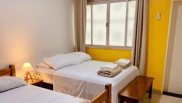 Une chambre avec deux lits, un banc, une table de chevet avec une lampe et une fenêtre avec des rideaux.