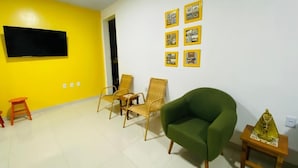 Une pièce avec un mur jaune, un téléviseur à écran plat fixé au mur et un fauteuil vert. On y trouve également deux chaises en osier et une petite table en bois sur laquelle est posée une statue.