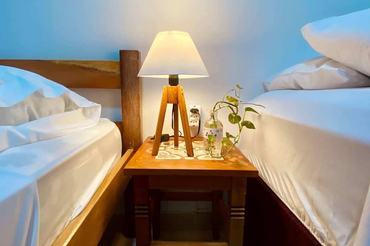 Quarto de hotel com duas camas, uma mesa de cabeceira de madeira com abajur e uma pequena mesa com um vaso de flores.