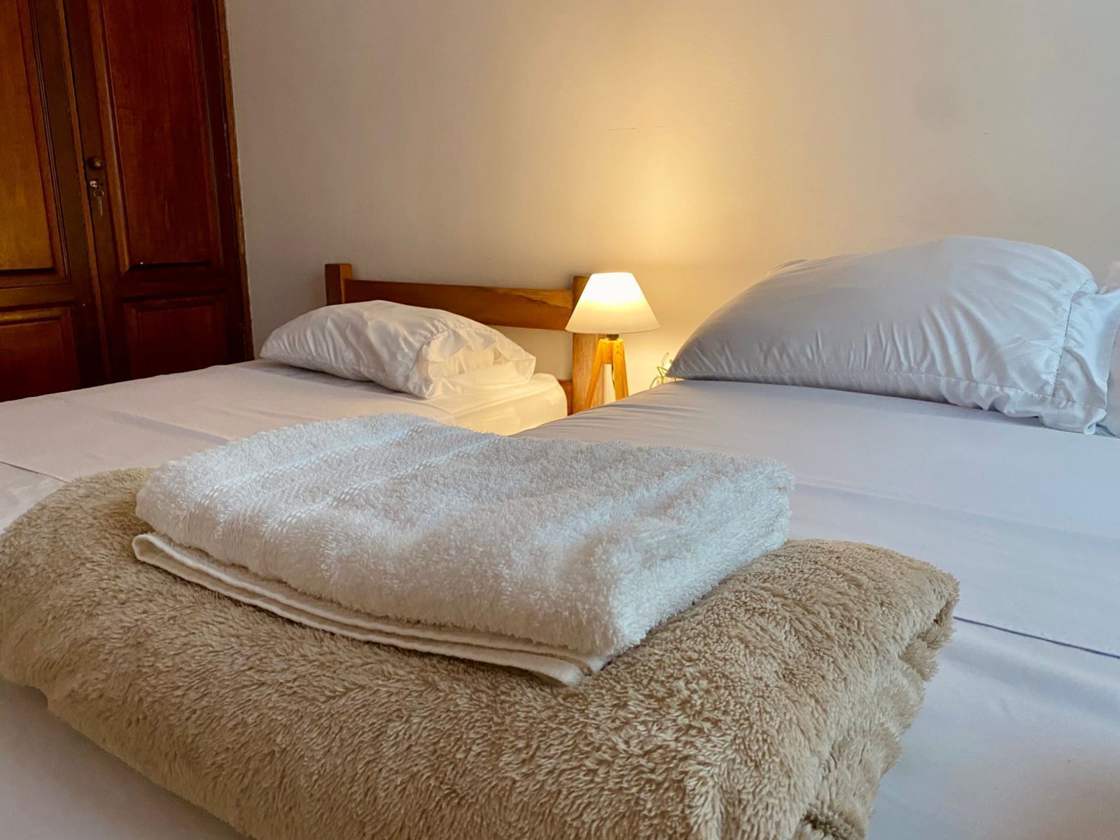 Cama bem arrumada com lençóis brancos, manta bege fofa e luminária de cabeceira.
