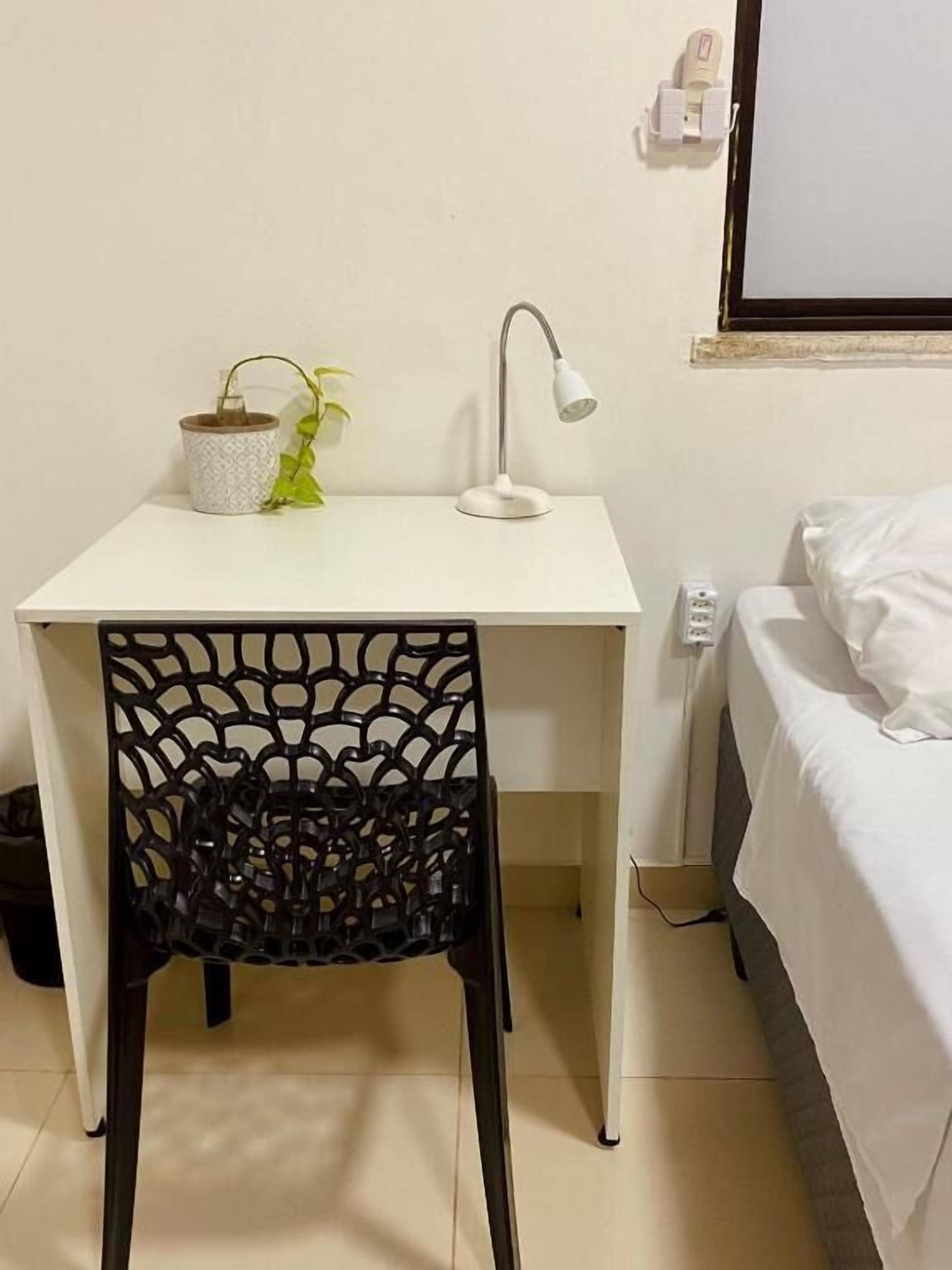 Un pequeño escritorio blanco con una lámpara y una planta en maceta. Hay una silla negra con respaldo de celosía. Se ve una cama con ropa blanca a la derecha.