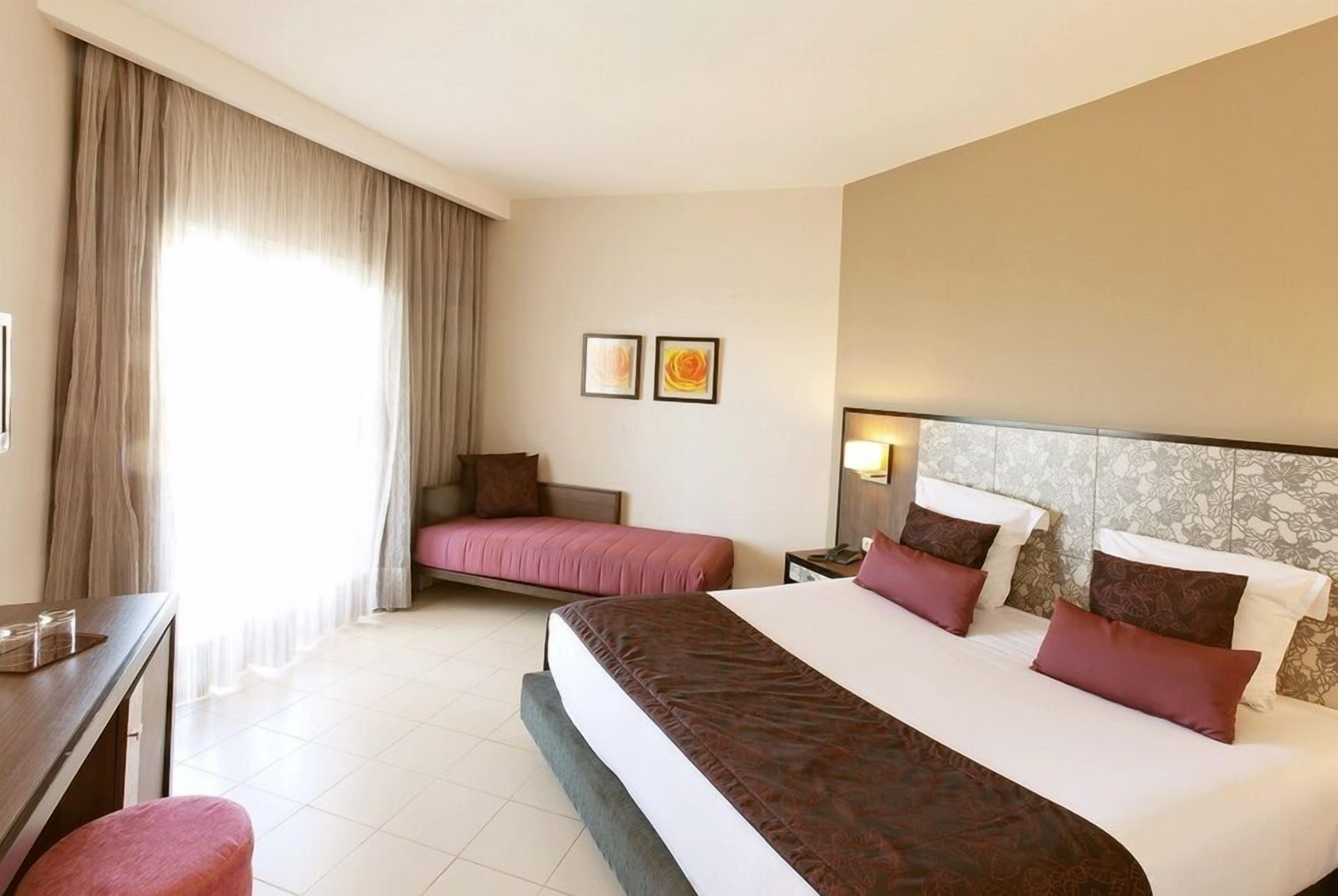A hotel room with a large bed, a smaller bed, a desk, and a chair. There are two paintings on the wall and a window with curtains.