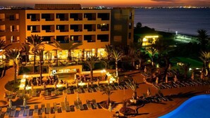 A hotel with multiple balconies overlooking a pool area, palm trees, and a view of the sea at dusk.