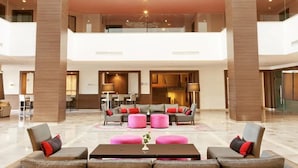 A spacious lobby with a high ceiling, a central seating area with sofas and ottomans, and a visible kitchen area in the background.