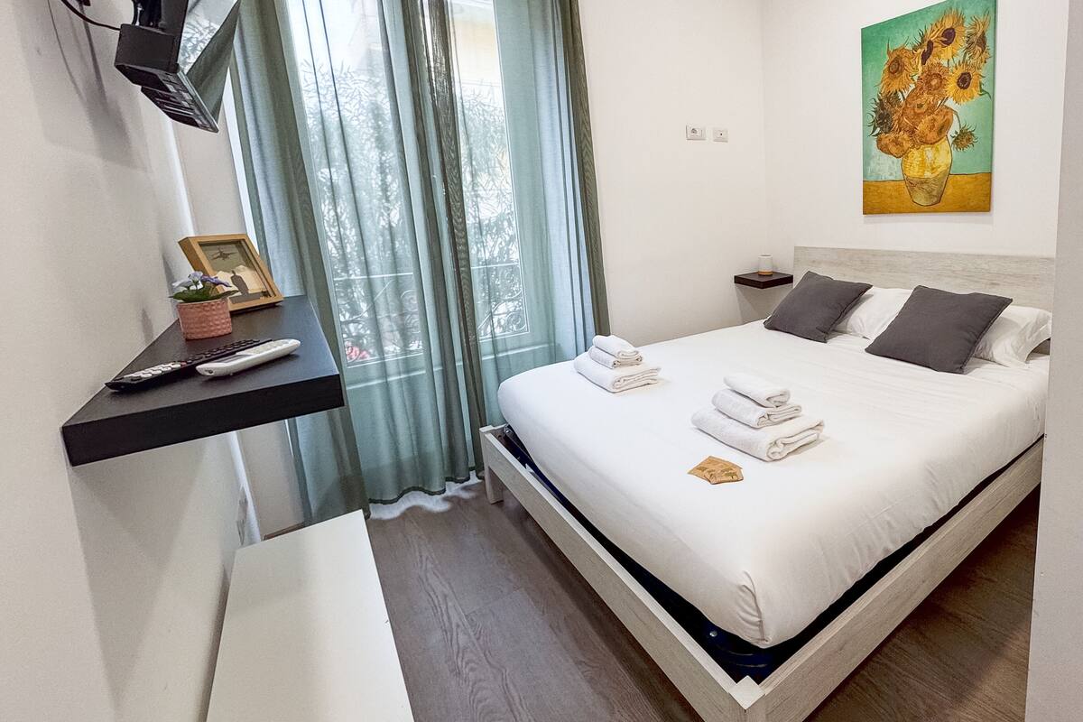 Ein Schlafzimmer mit Bett, Nachttisch, Lampe, Vorhängen und einem Gemälde.