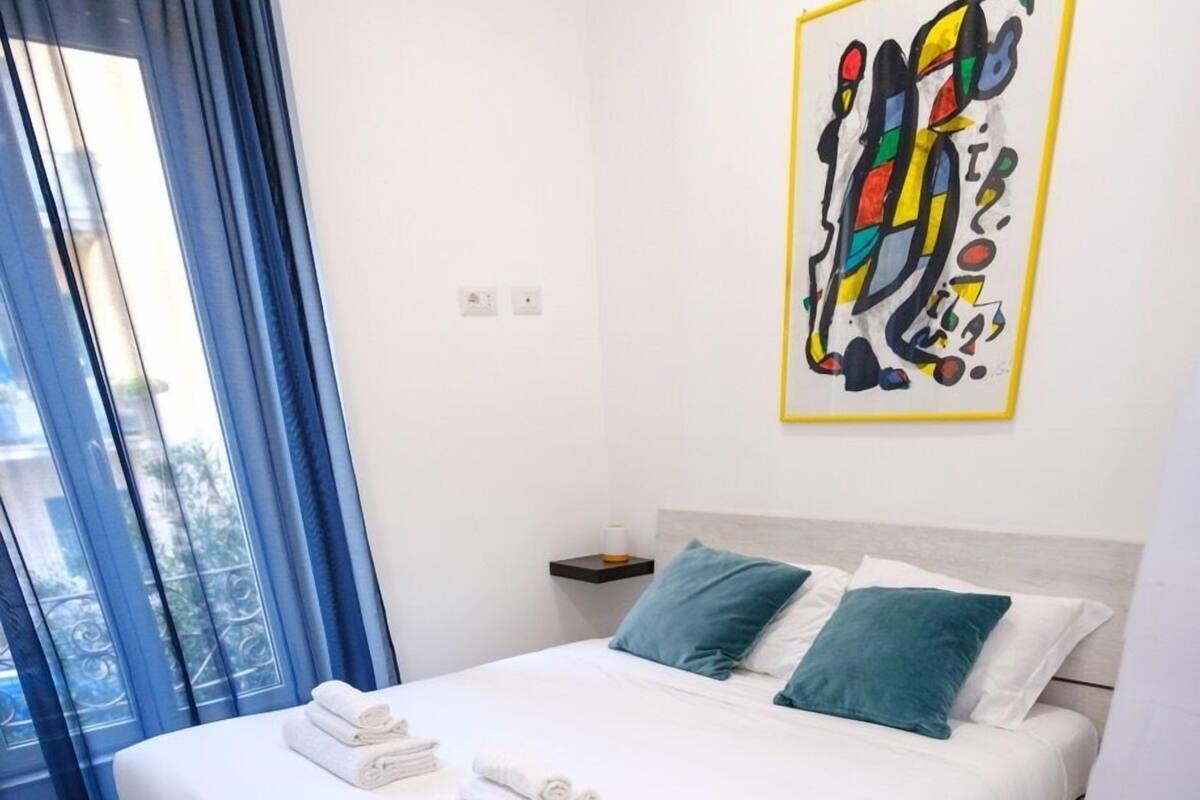 Ein ordentlich bezogenes Bett mit weißen Leinen und gefalteten Handtüchern, ein Nachttisch mit Lampe sowie ein farbenfrohes abstraktes Gemälde an der Wand.