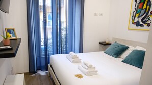 A neatly made bed with white linens and teal pillows, a small bedside table with a lamp, and a window with blue curtains. - Denina67 - ChiacchioProperty (Rome)