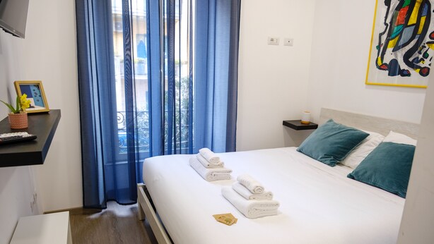 A neatly made bed with white linens and teal pillows, a small bedside table with a lamp, and a window with blue curtains. - Denina67 - ChiacchioProperty (Rome)