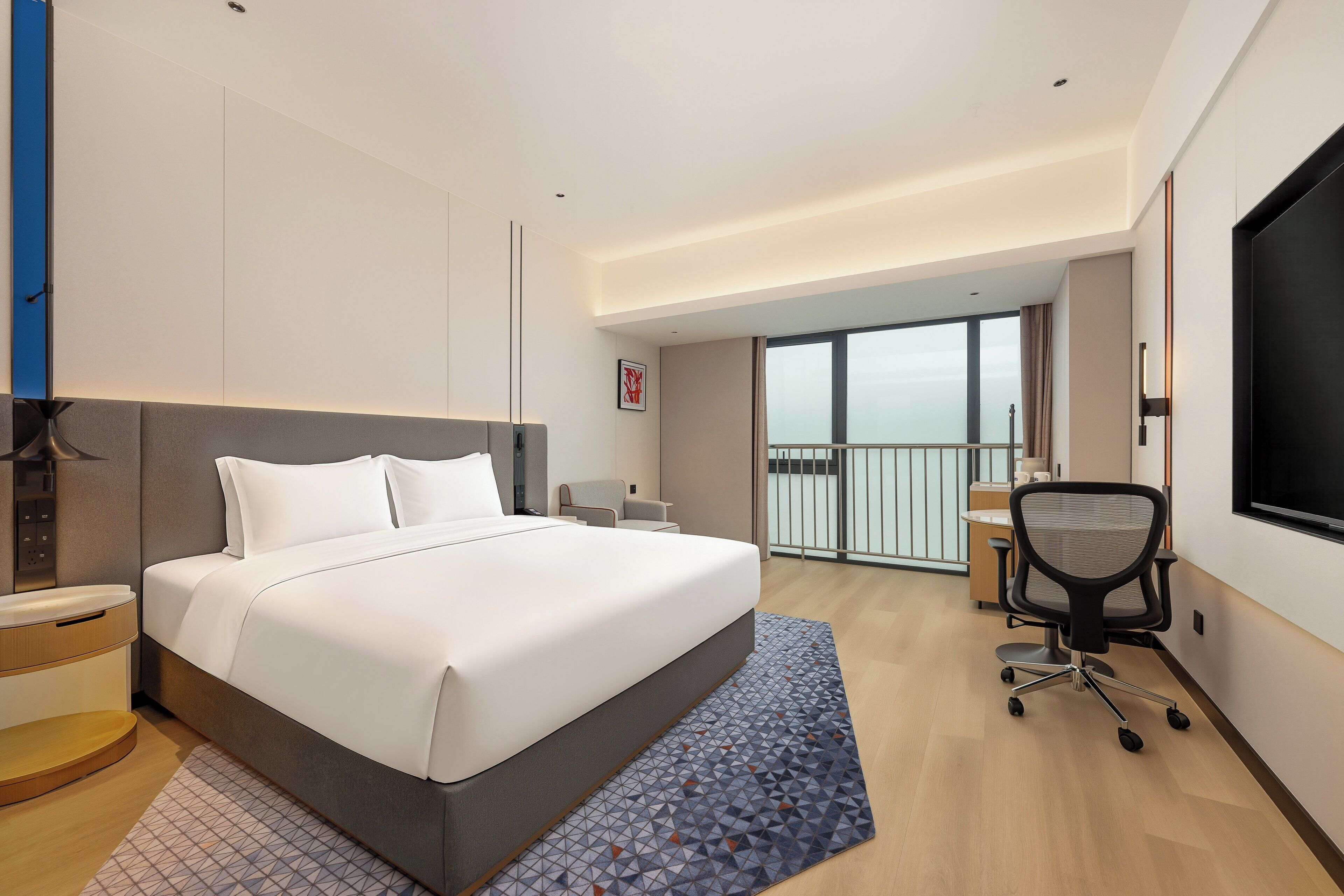 A modern hotel room with a large bed, a desk, and a view of the city.