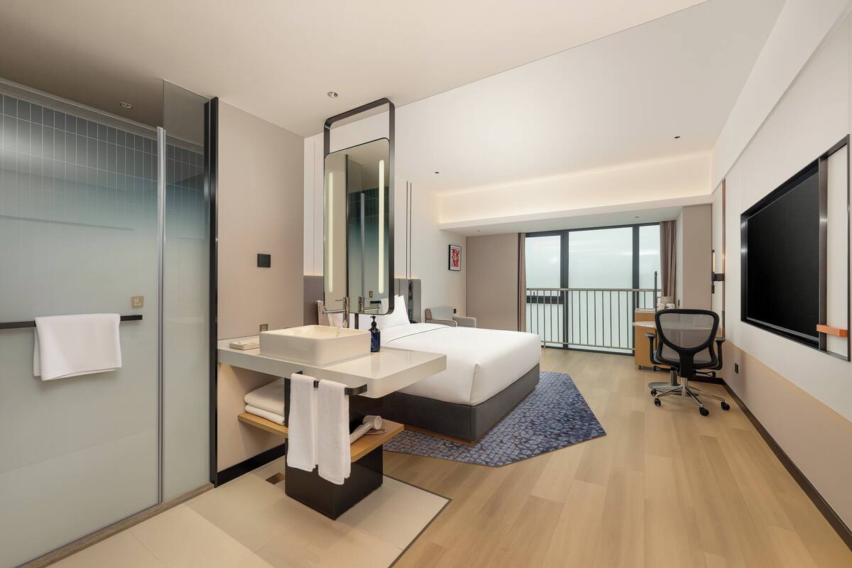 Camera d'albergo moderna con un letto grande, bagno con box doccia in vetro, una scrivania e un'ampia finestra vista panoramica.