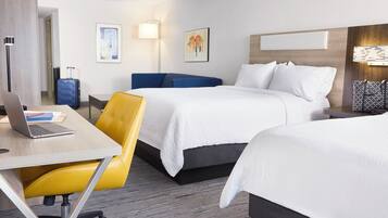 A hotel room with two beds, a desk with a laptop, a chair, a suitcase, and artwork on the walls.