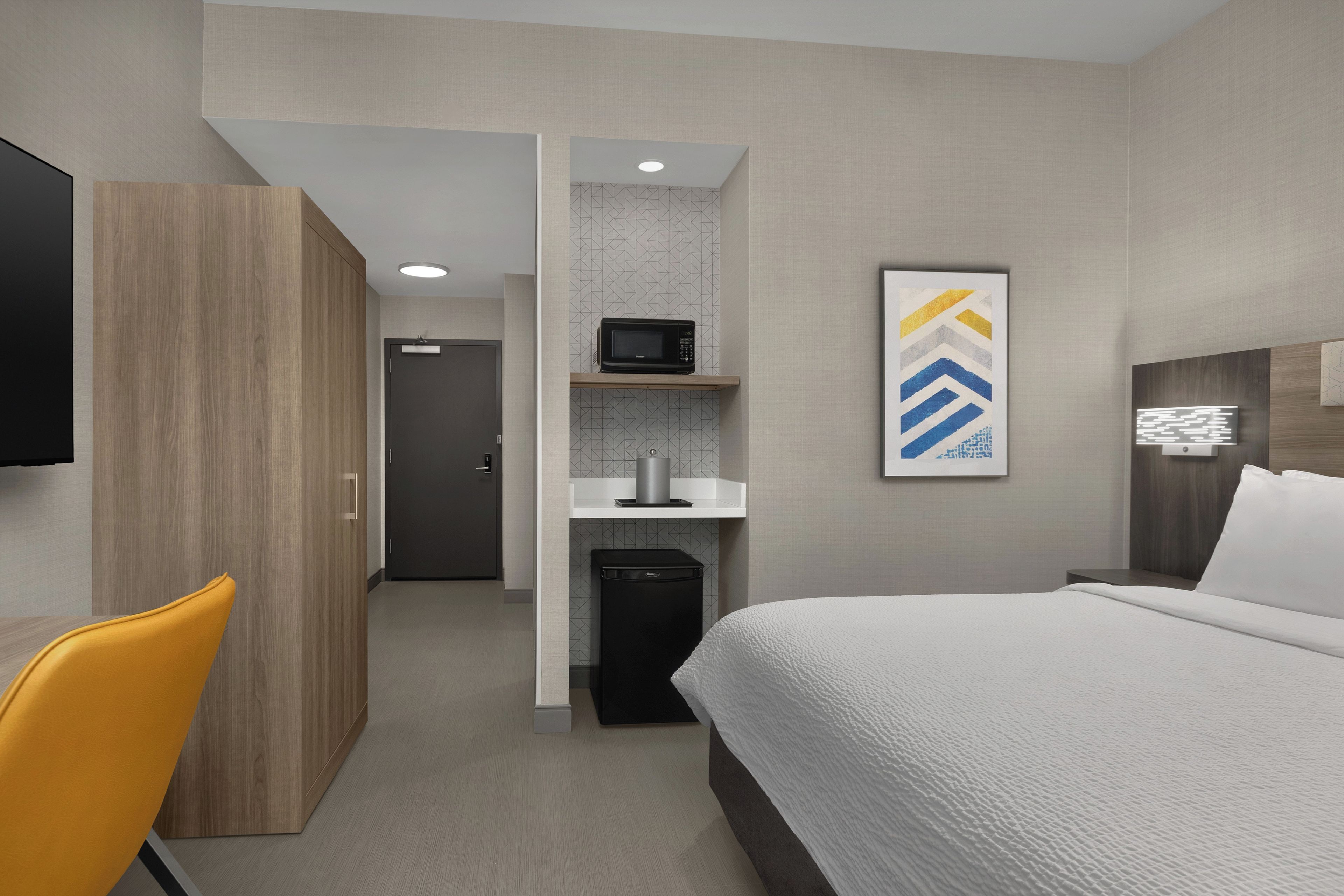 Habitación de hotel con cama, escritorio, silla, microondas y una pequeña kitchenette.