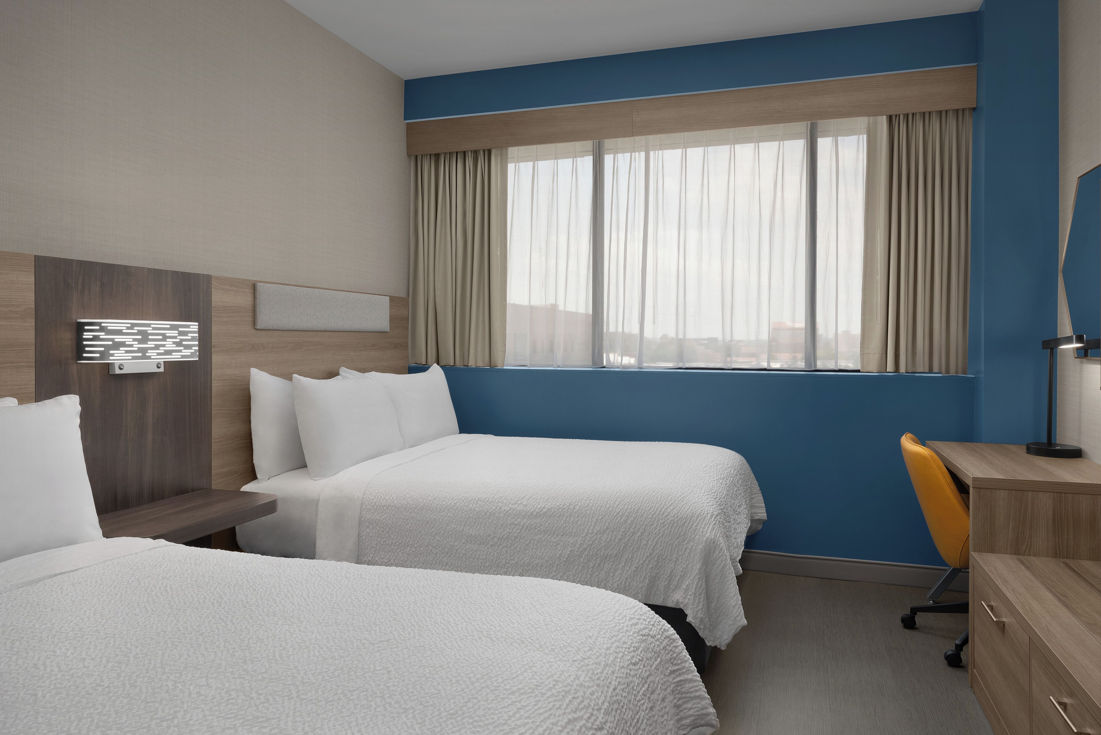 A hotel room with two beds, a desk, a chair, and a large window with curtains.