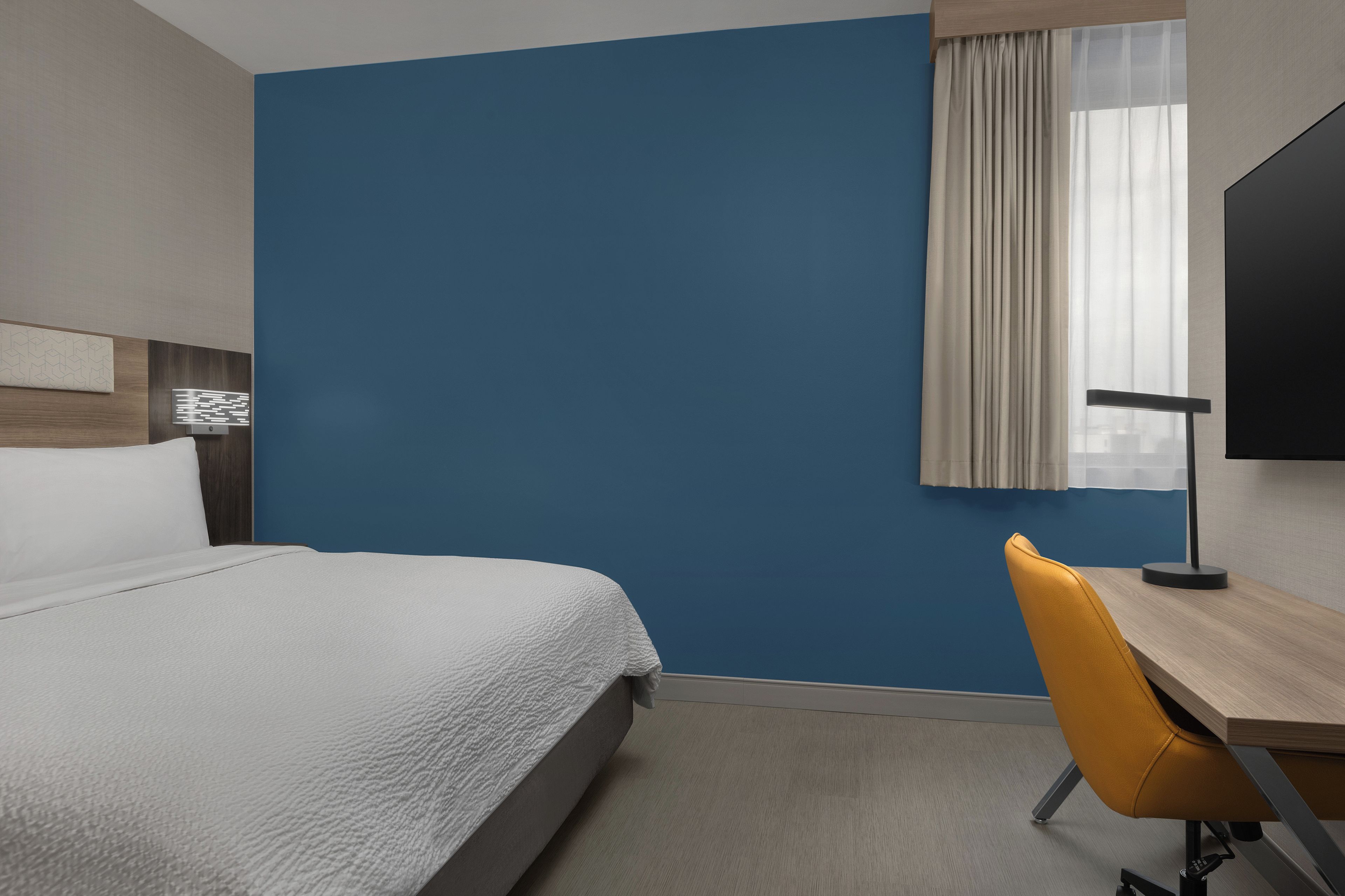 Habitación de hotel con cama, escritorio, silla, televisor y ventana con cortinas.