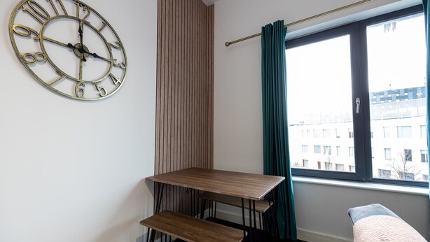 A room with a wooden table, a bench, a wall clock, and a window with curtains. - Spacious 2 bed apt Near Media City and Old Trafford (Salford)