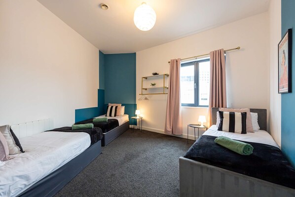 A room with two beds, a window with curtains, a small table with a lamp, and a wall-mounted shelf with decorative items. - Spacious 2 bed apt Near Media City and Old Trafford (Salford)