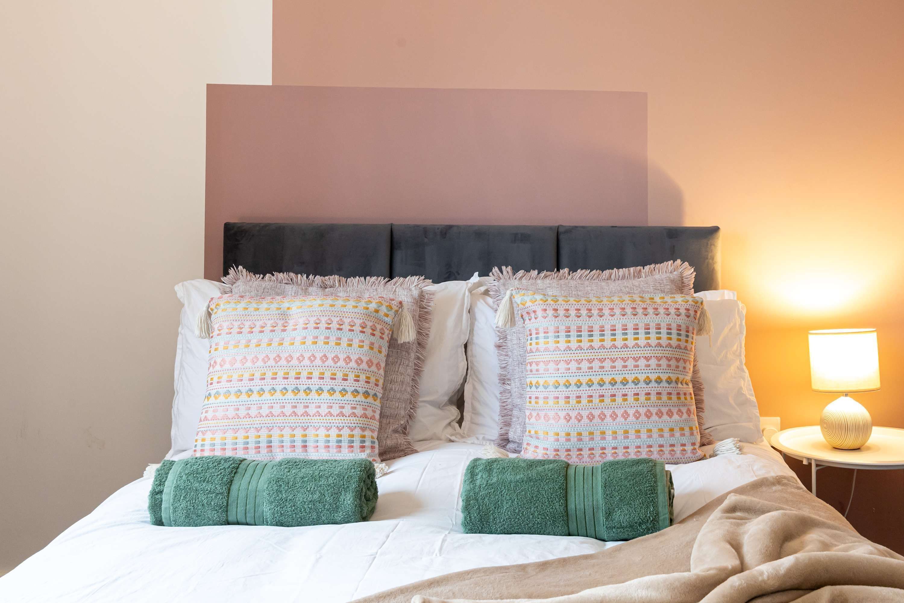 A neatly made bed with patterned pillows and green towels, a bedside lamp, and a plain wall.